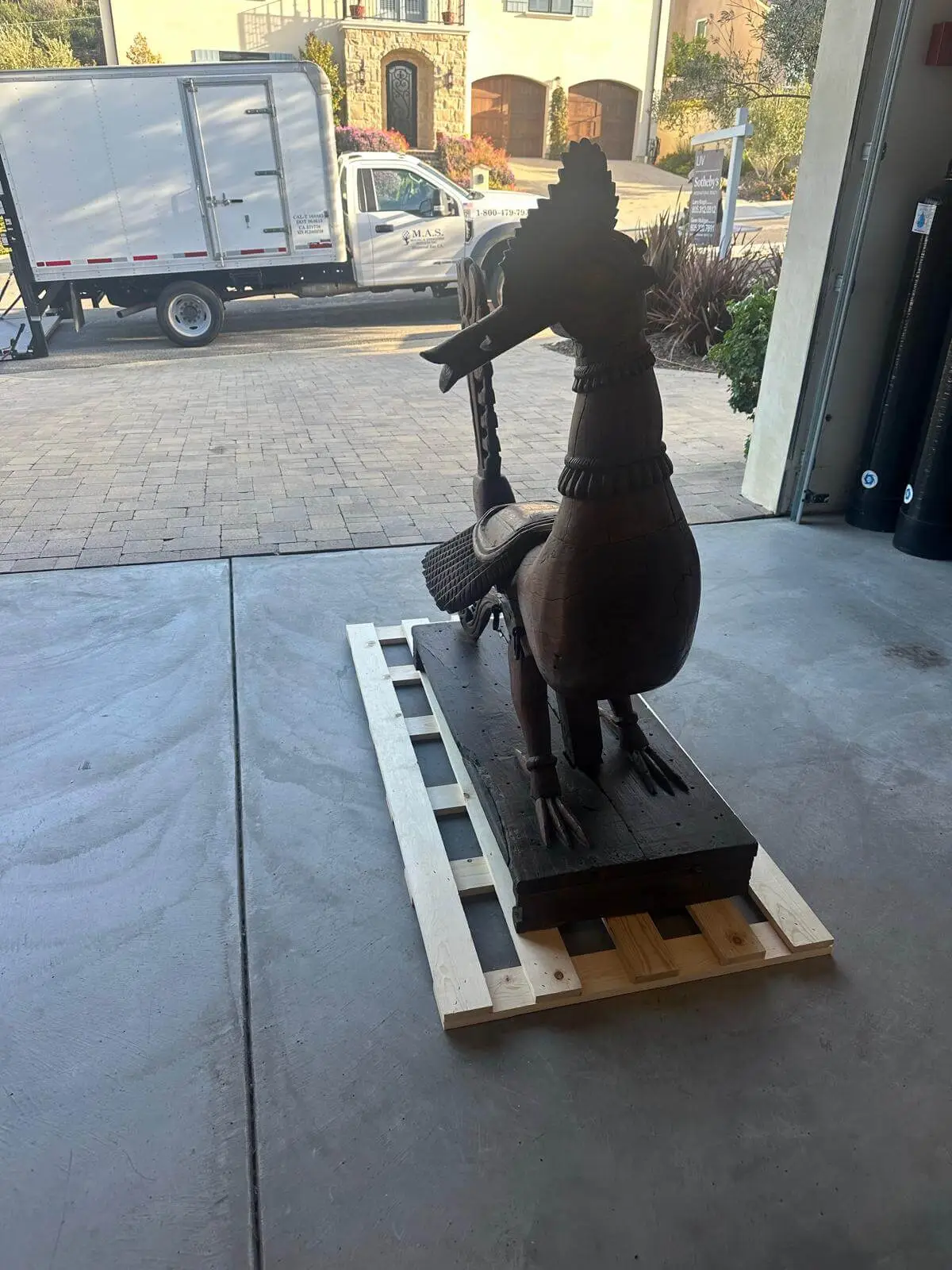 A carved wooden bird sculpture fixed to a pallet base in the driveway before crating.