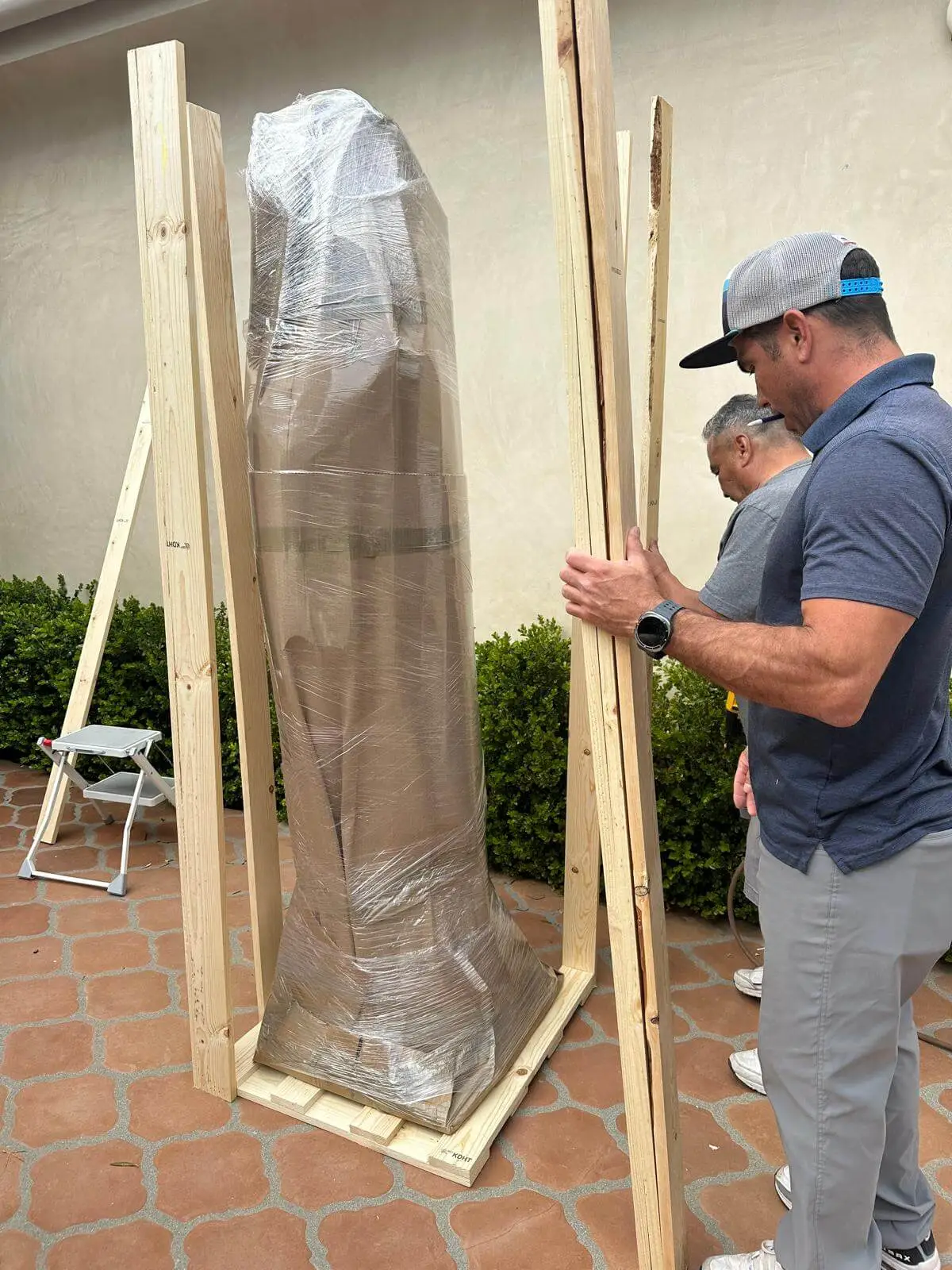 Two crew members standing a tall wrapped piece into its crate frame on a patio.