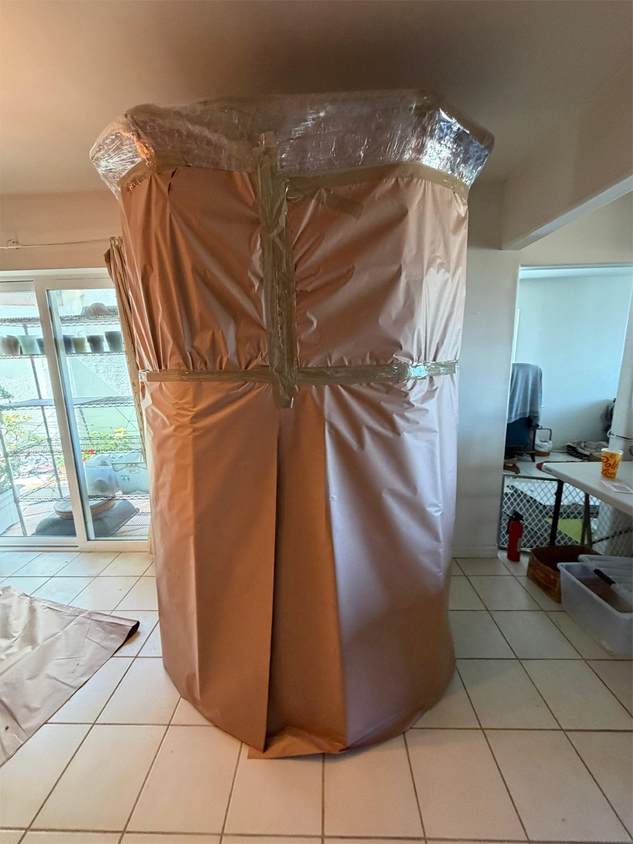 A tall ceramic urn wrapped in kraft paper and banded with stretch film, standing ready in a tiled entryway.
